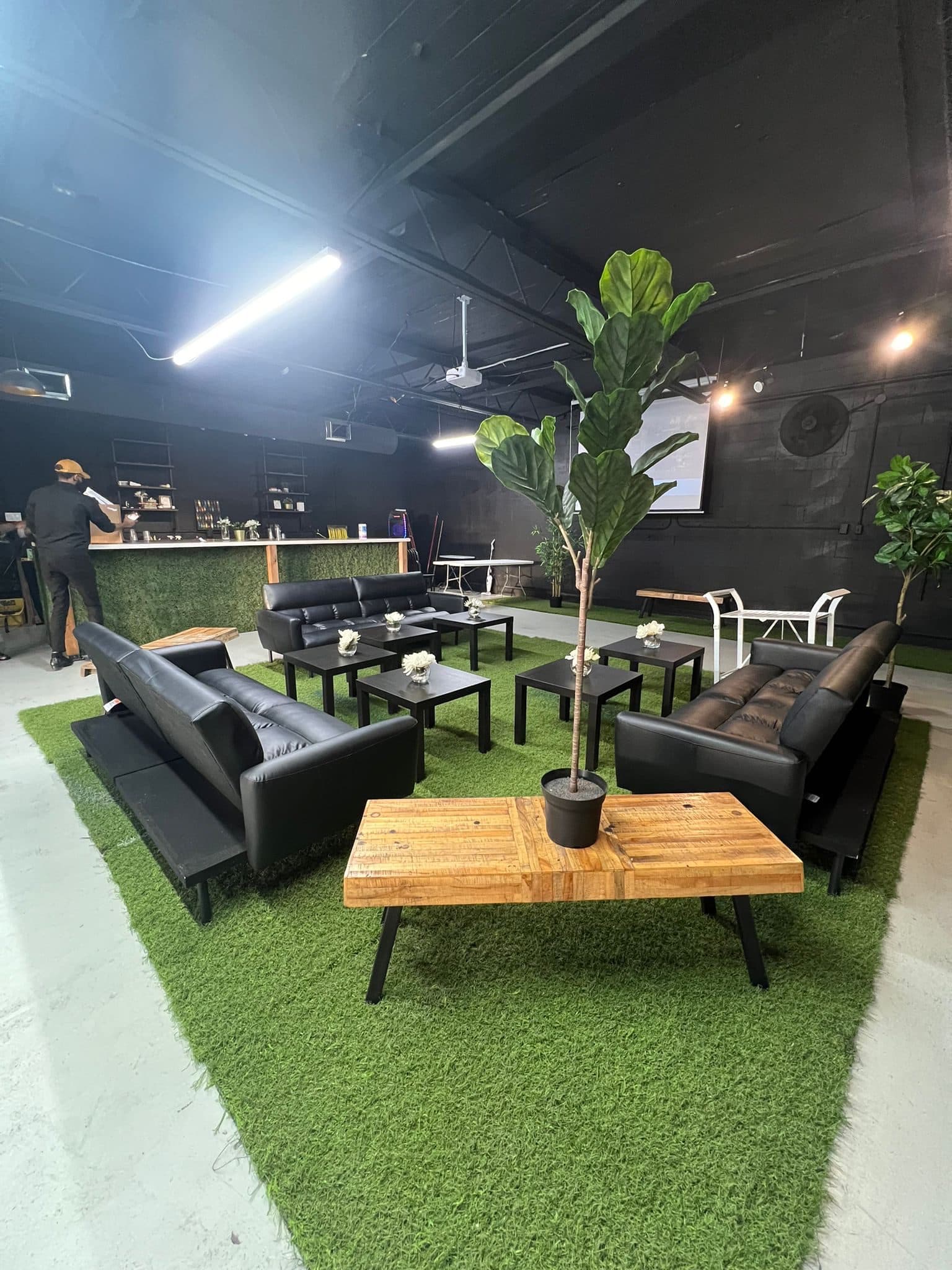 A lounge arrangement at Half Note: black leather sofas and coffee tables on turf, potted plants, and a projector screen on the wall.