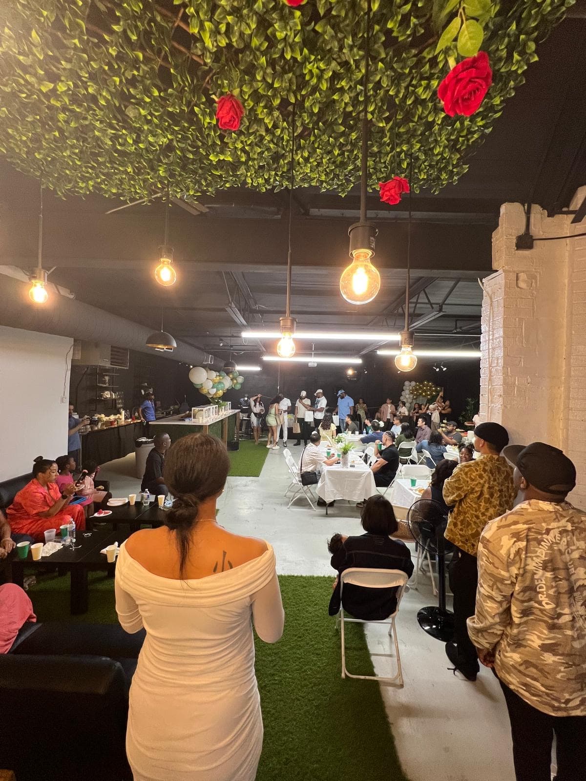 Guests at a party at Half Note, seated at white-linen tables and lounge sofas under a ceiling of greenery and roses with hanging bulbs.