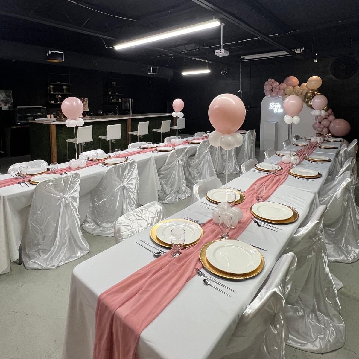 A baby shower set up at Half Note: long banquet tables with white linen, gold chargers, a blush runner and a balloon arch.