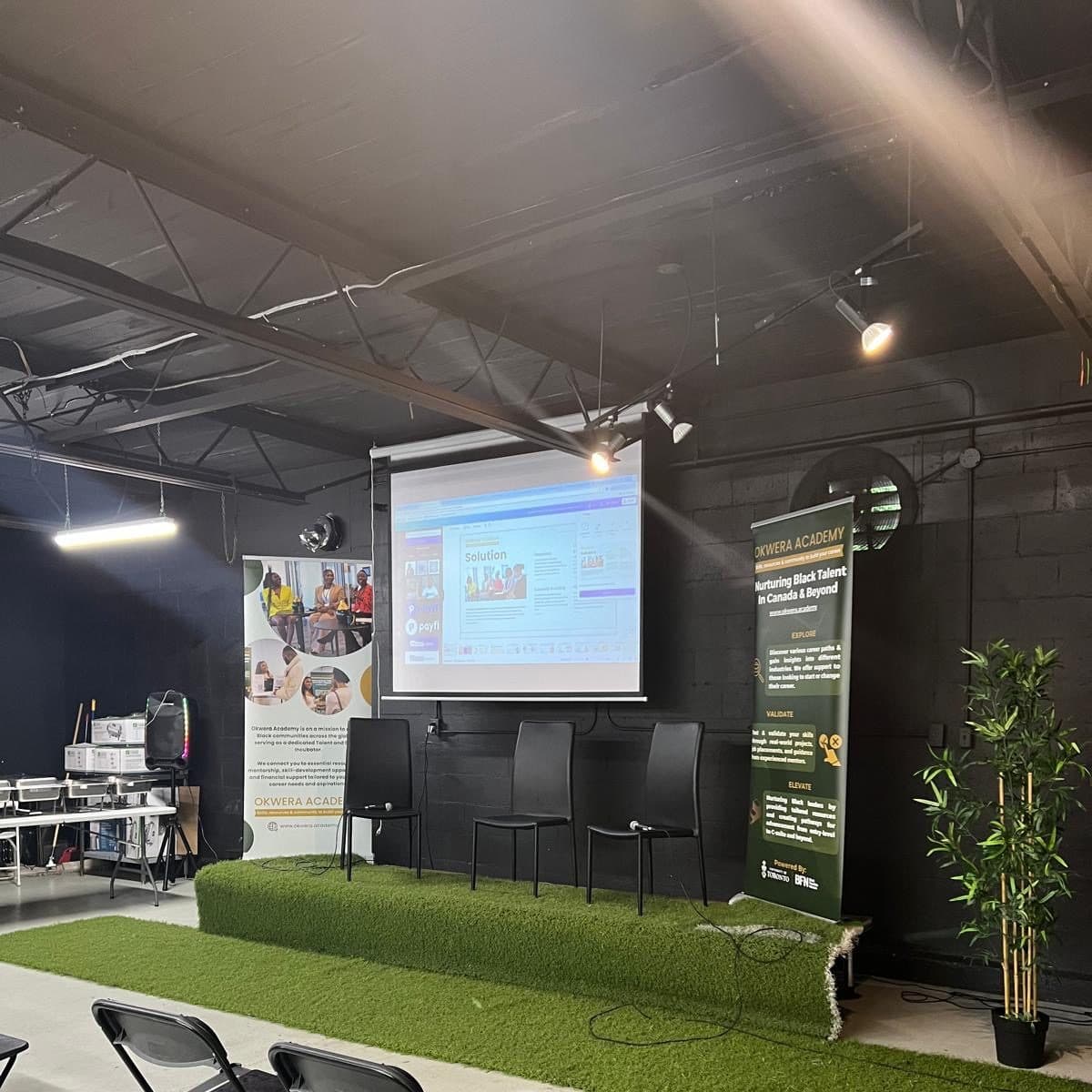 A panel set up at Half Note: three chairs on a turf-covered riser in front of a projector screen, with pull-up banners either side.